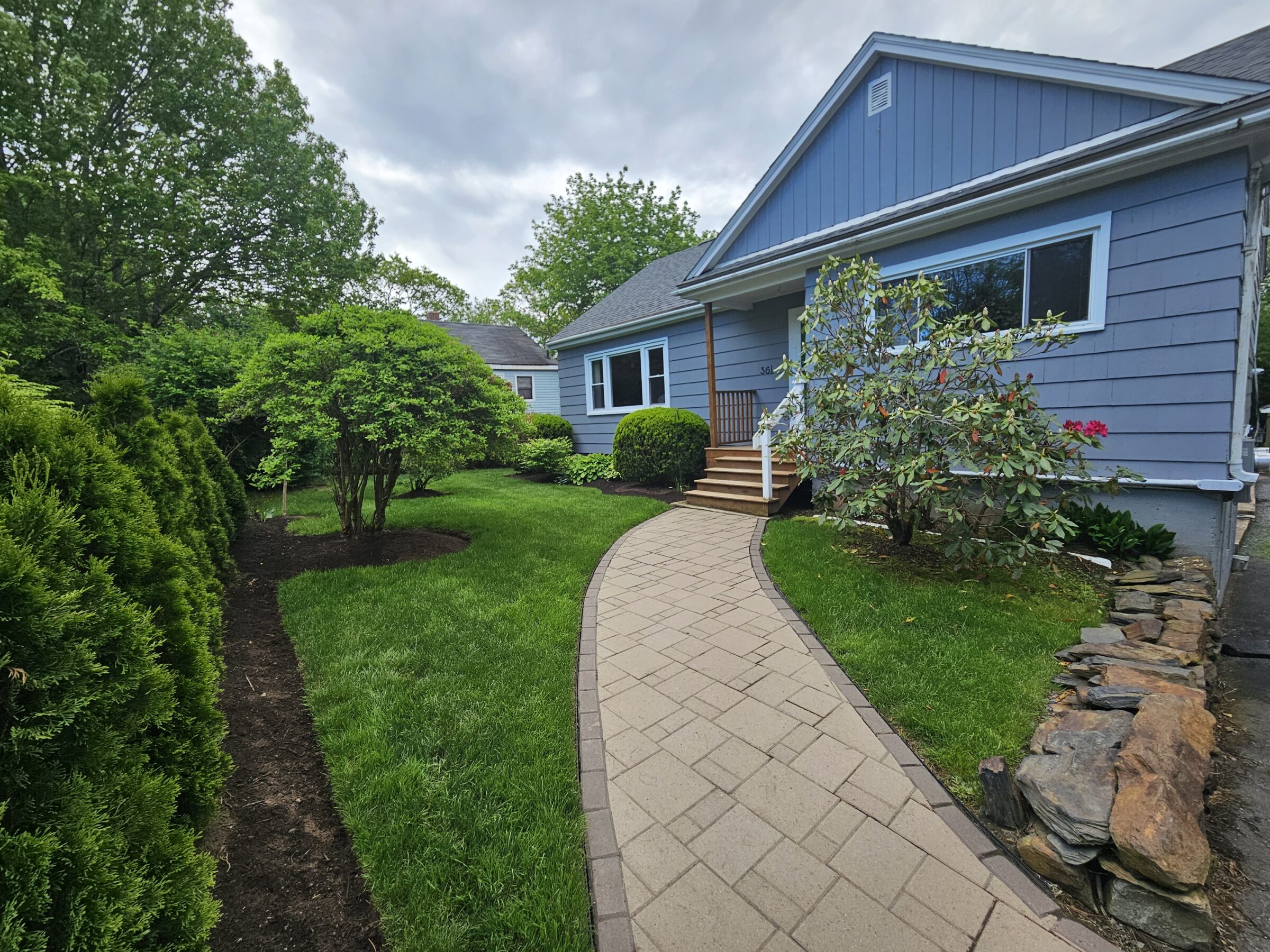 A blue house with lush green landscaping, a stone pathway, and flowering shrubs. No people or recognizable landmarks are visible.