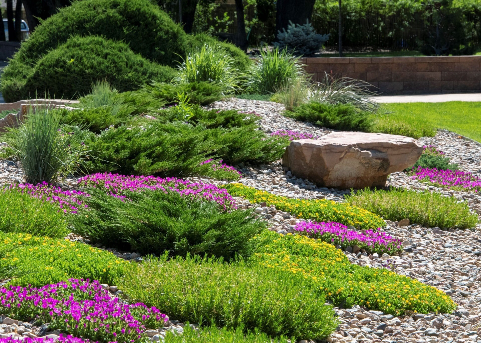 A beautifully landscaped garden features vibrant purple and yellow flowers, green shrubs, and decorative rocks, surrounded by lush greenery under a clear sky.