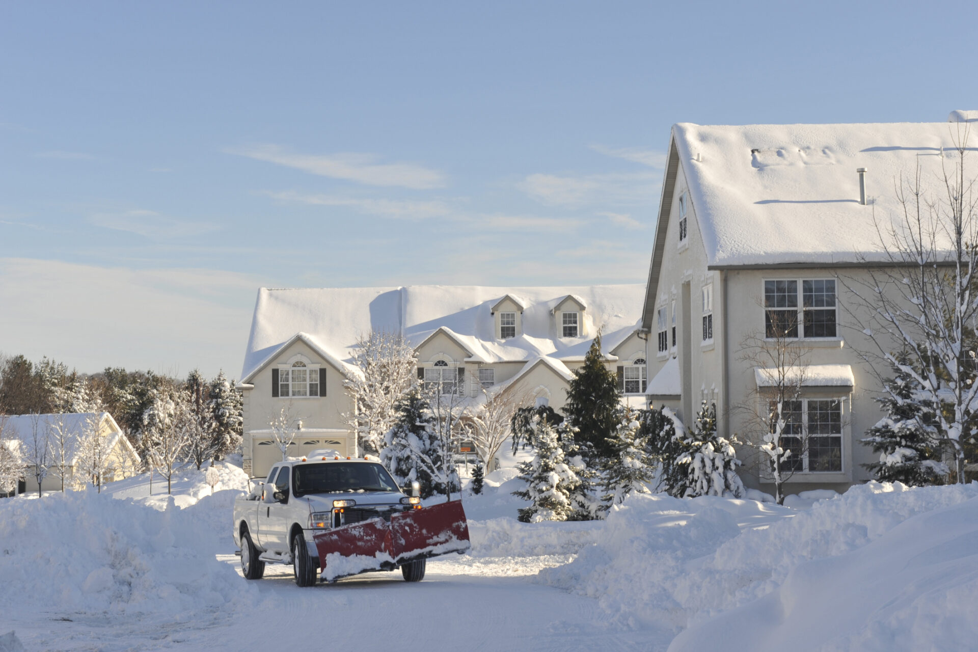 A snow-covered suburban street with houses. A truck with a plow is clearing snow under a clear blue sky, surrounded by trees.