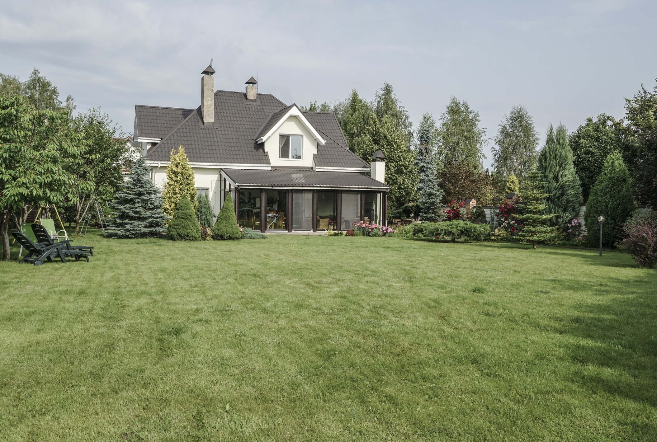 Charming suburban house with dark roof, large green lawn, and lush garden. Two lounge chairs are placed near trees in the backyard.