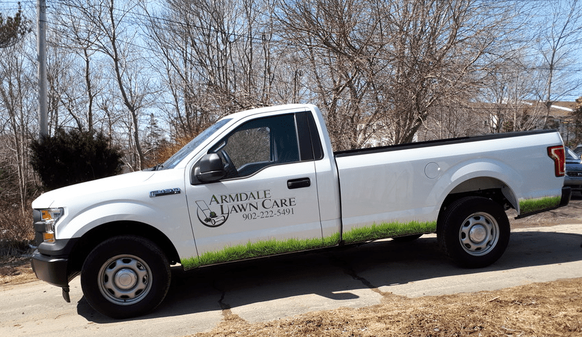 White pickup truck with "Armdale Lawn Care" logo and contact number parked on a street. Background features leafless trees and a clear sky.