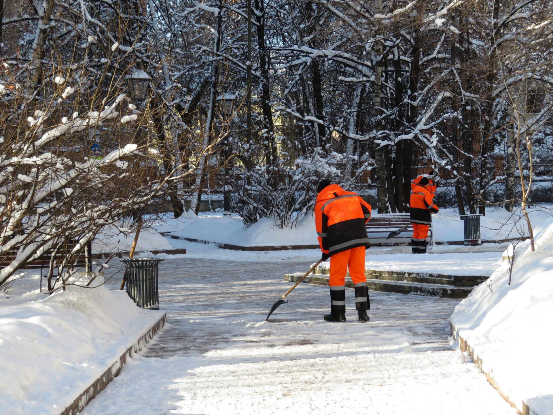 Two persons in orange jackets clear snow from park pathways surrounded by snow-covered trees and lampposts. Trash bins line the path.