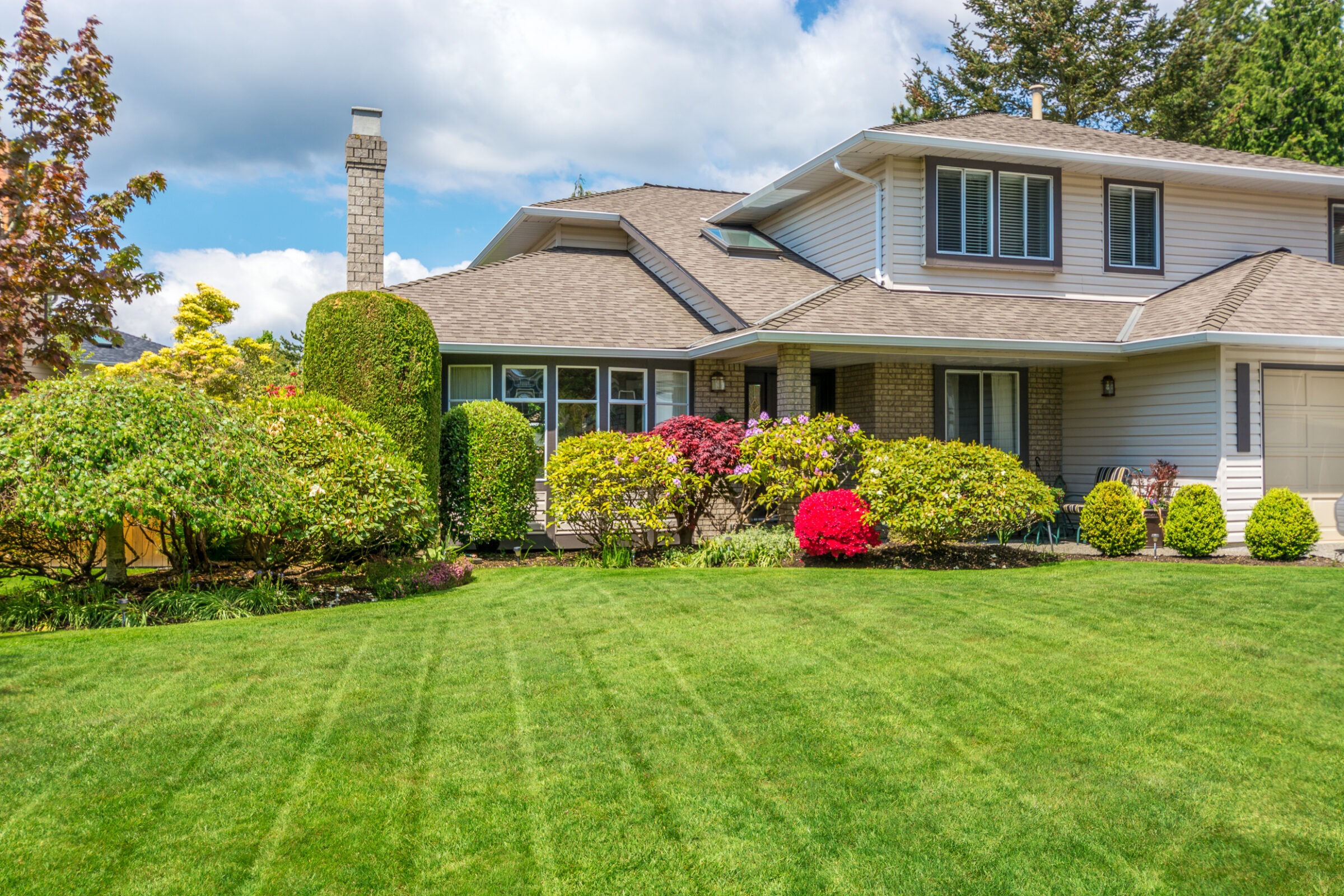 A suburban house with a manicured lawn and colorful shrubs under a partly cloudy sky, showcasing classic architecture and pleasant landscaping.