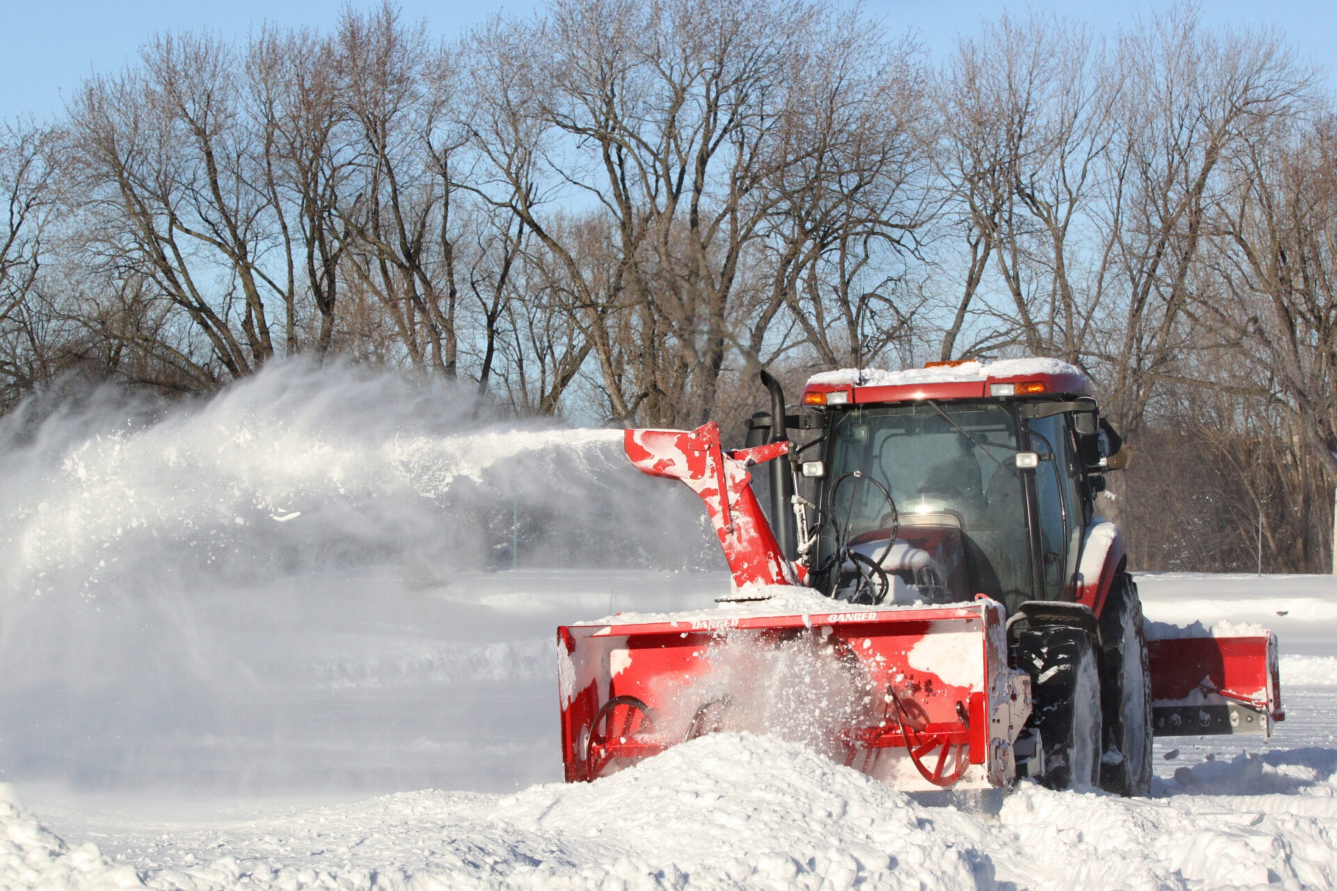 A red snowplow removes snow in a winter landscape with bare trees, operated by a person inside the plow.