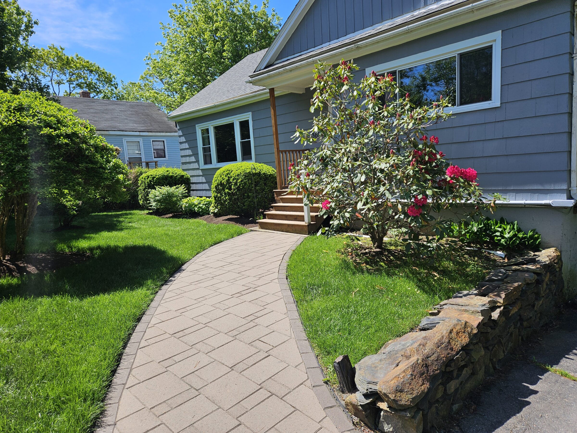 A quaint house with a well-maintained pathway, lush green lawn, and blooming shrubbery on a sunny day. No recognizable landmarks or buildings.