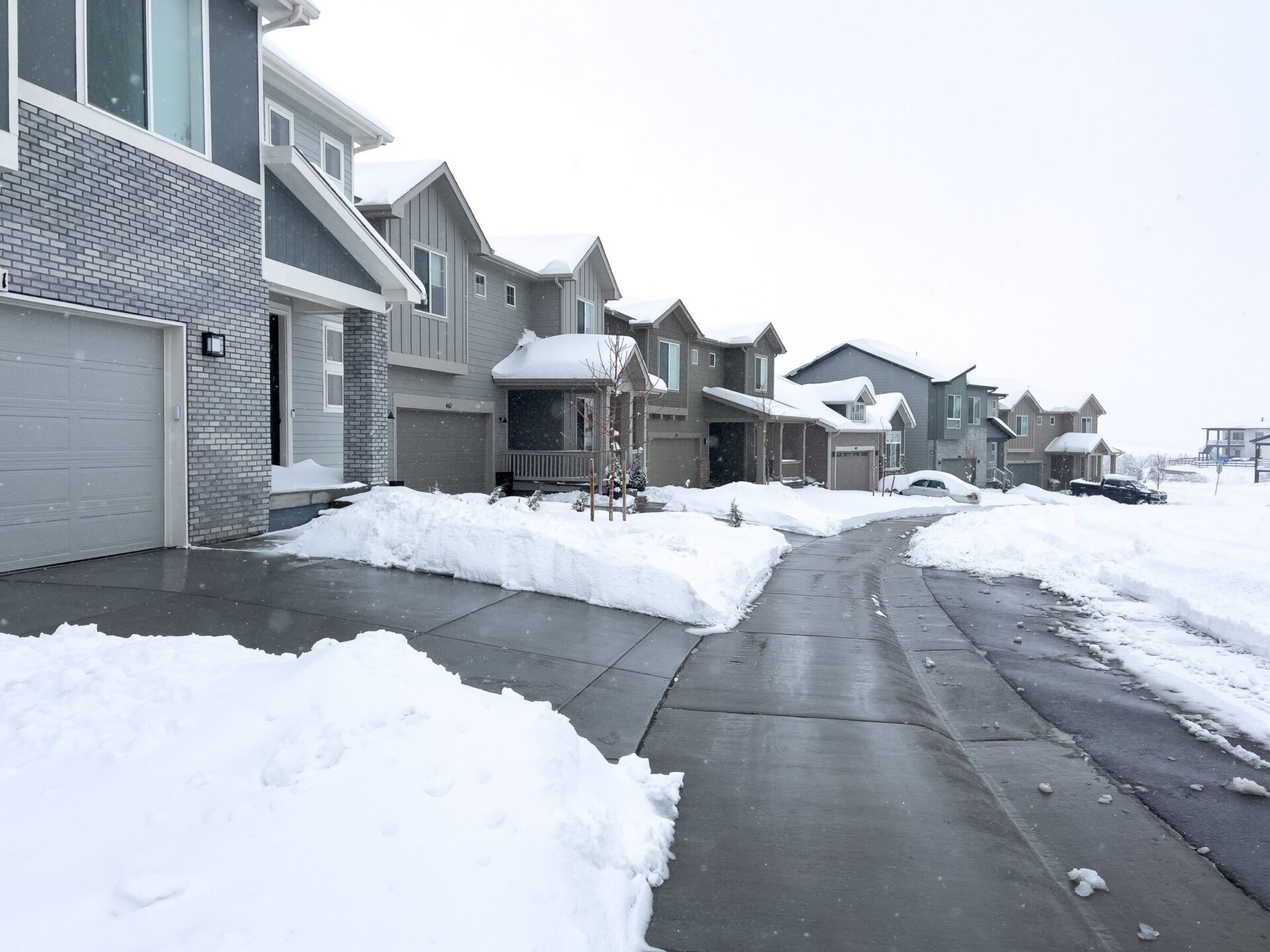 Snow-covered suburban street with modern houses, clear driveways, and parked cars. Overcast sky adds to the winter atmosphere.