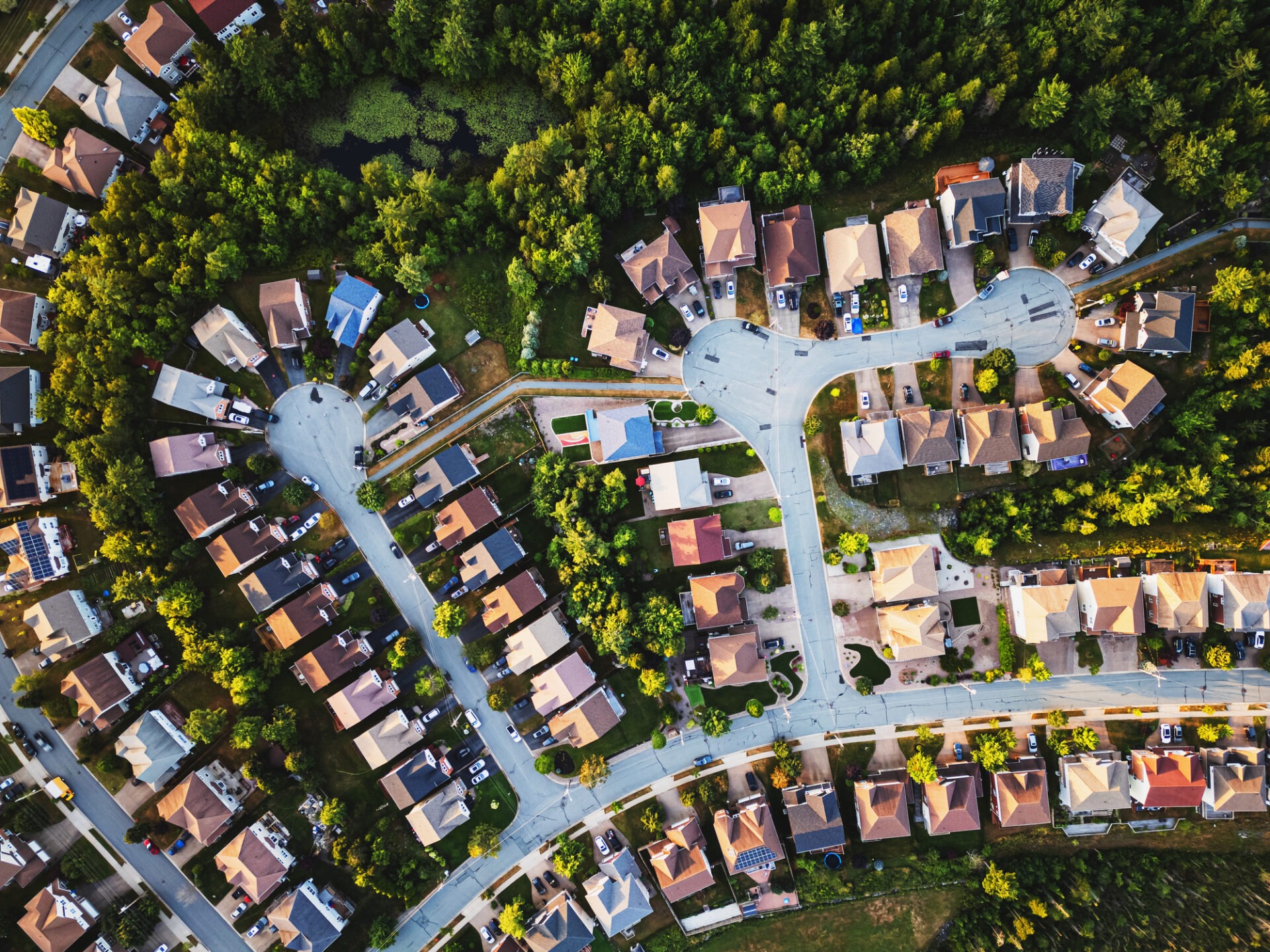 Aerial view of a suburban neighborhood with curved streets, numerous houses, parked cars, and surrounding greenery, showcasing a typical residential layout.