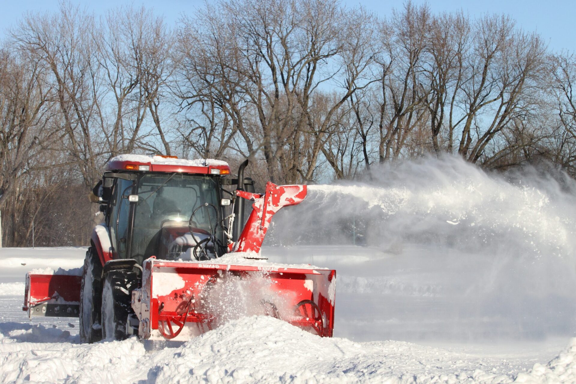 A red snowplow clears snow in a winter landscape, with bare trees in the background and clear blue skies overhead.