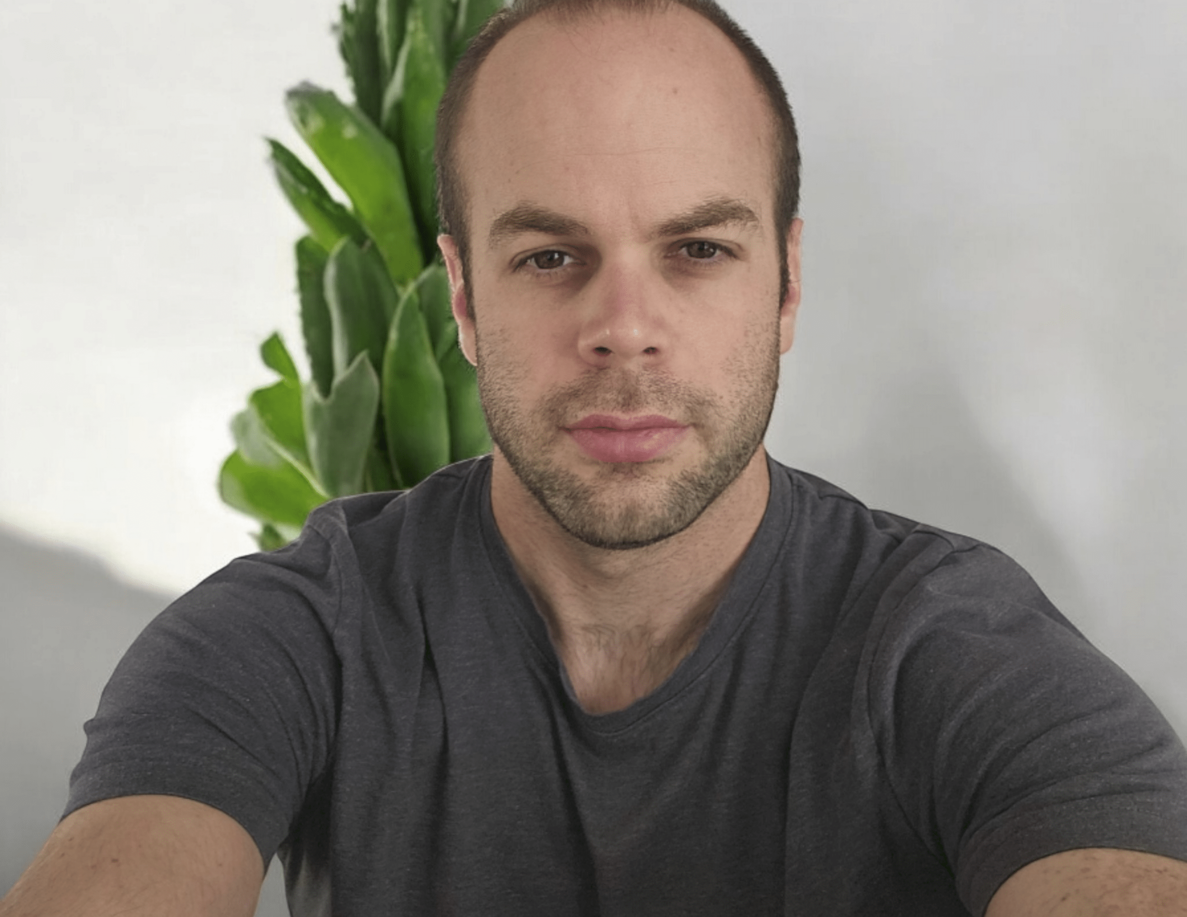 A person in a gray shirt is taking a selfie in front of a green plant, with a neutral background.