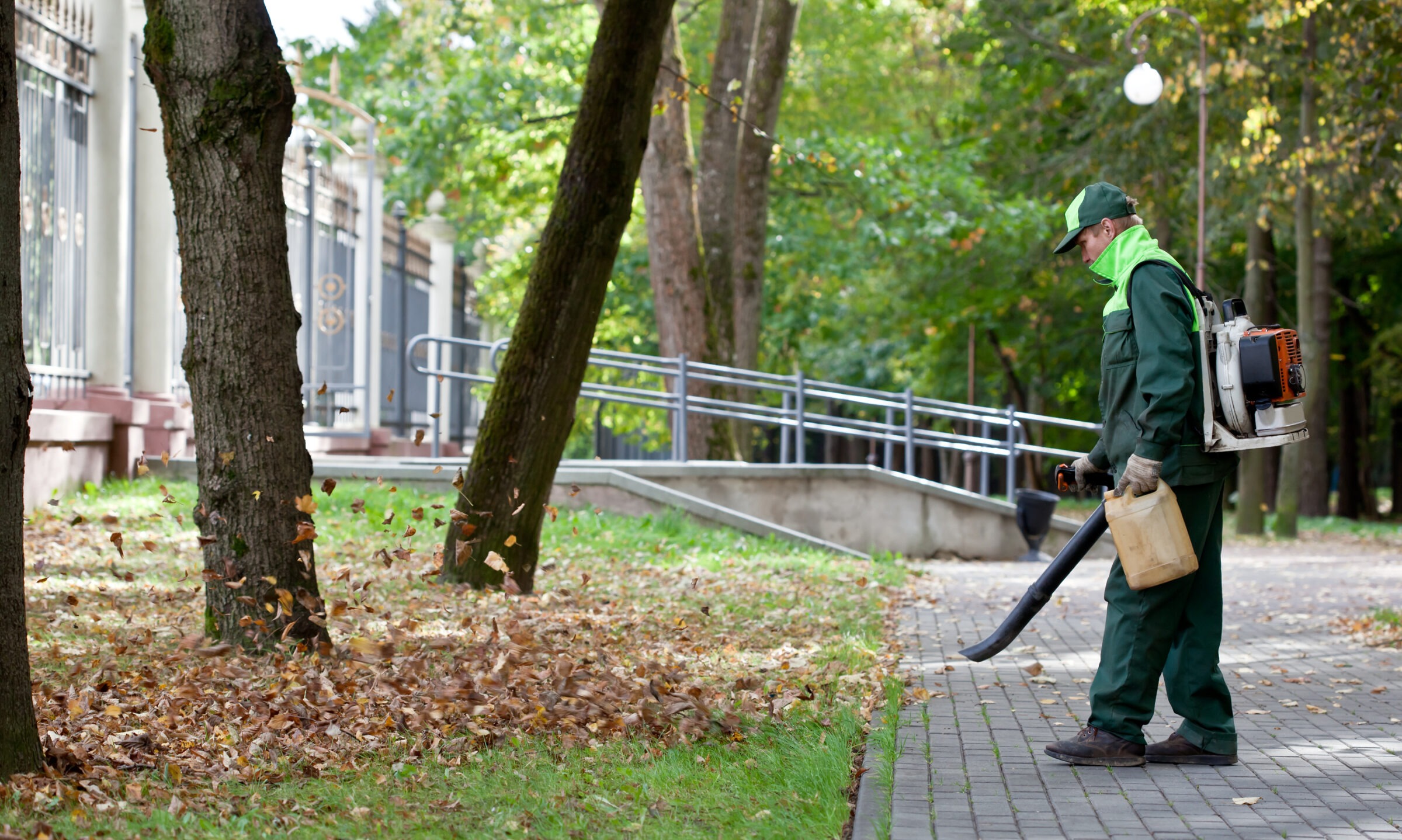 A person in green uniform uses a leaf blower on a tree-lined pathway next to a metal fence in a park setting.