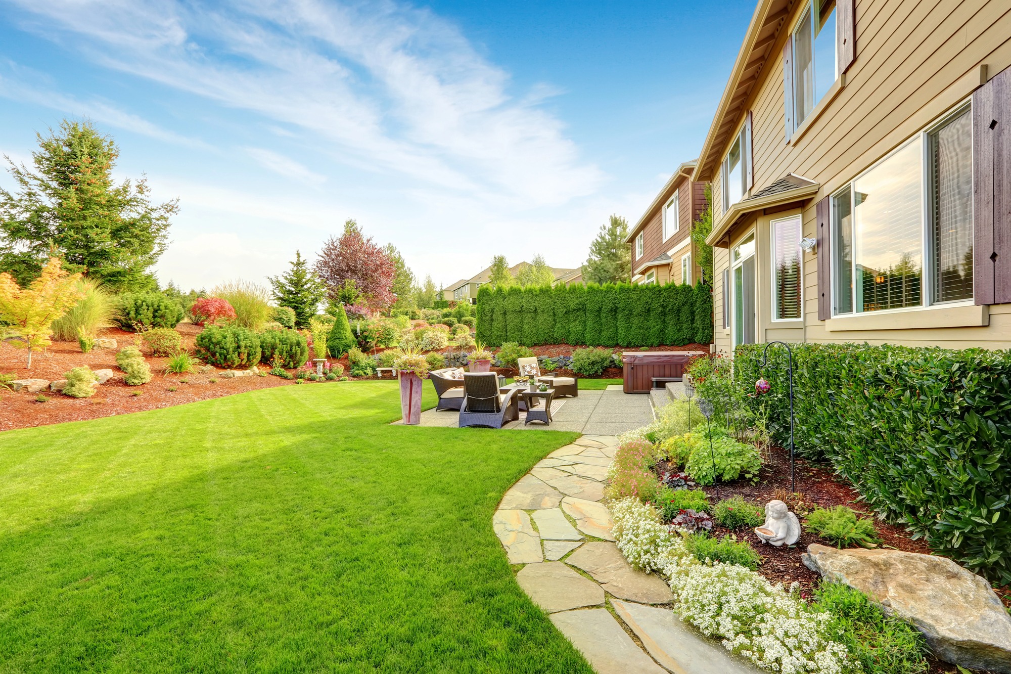 A beautifully landscaped backyard with lush green lawn, stone pathway, patio seating, and vibrant garden, adjacent to a modern house under clear skies.