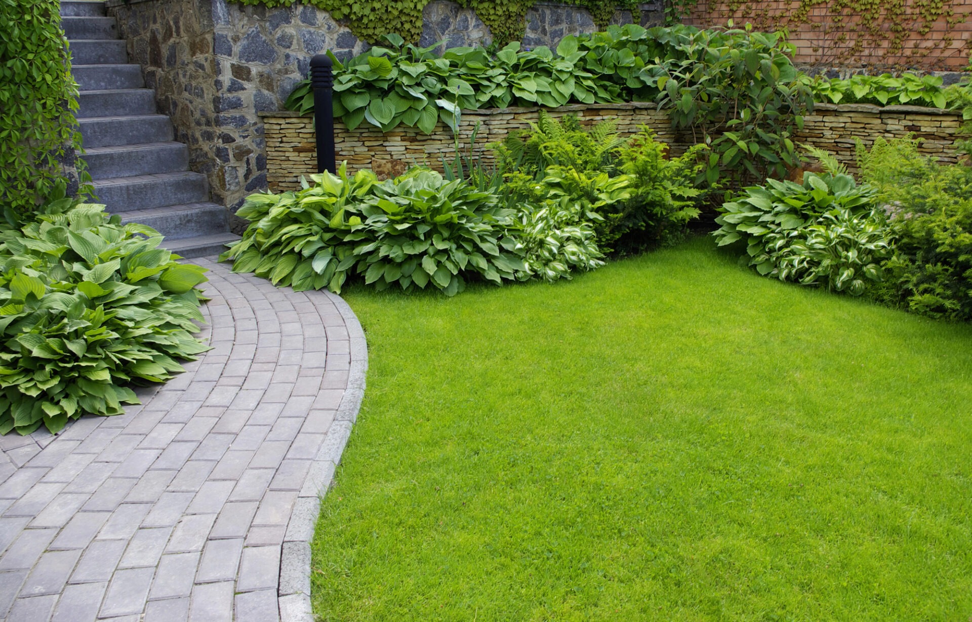 A beautifully landscaped garden featuring lush greenery, neatly trimmed grass, stone pathway, and steps against a stone wall covered in climbing vines.